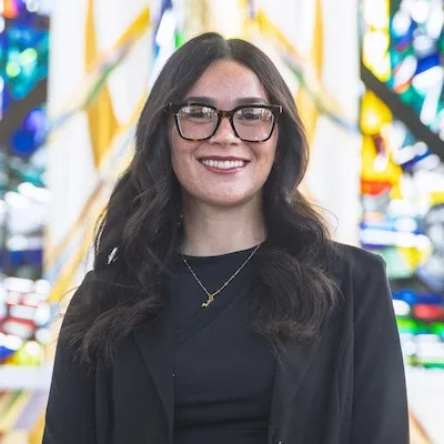 Valparaiso University student with long dark hair, glasses, and a black blazer smiles in front of a colorful stained glass window in the Chapel of the Resurrection on campus.
