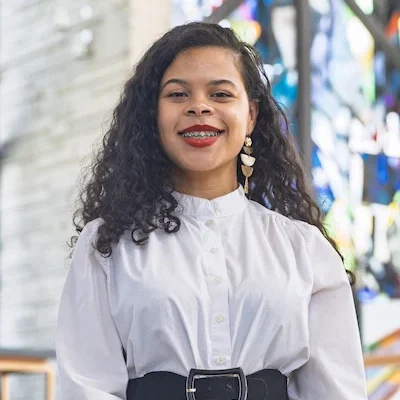 Valparaiso University student in a white dress with a black belt, smiling, standing with colorful stained glass in the background in the Chapel of the Resurrection on campus.