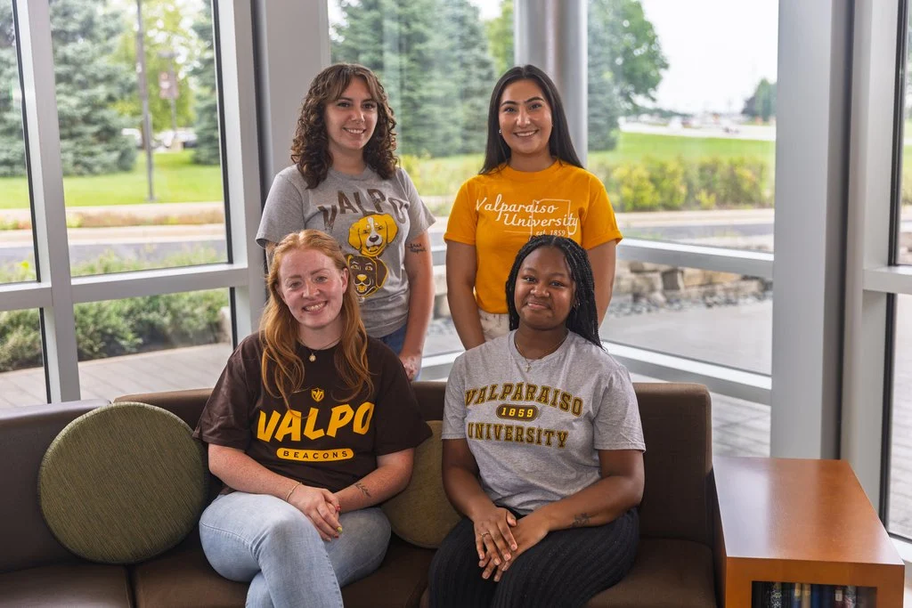 Four students in Valparaiso University shirts smile indoors by large windows with greenery outside, enjoying a visit with Admission Counselors.