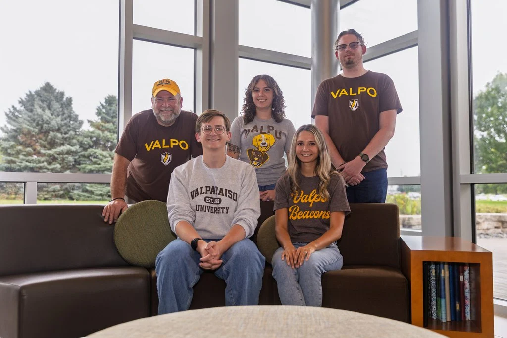 Five Valparaiso University admission counselors wearing Valpo shirts pose together indoors, sitting and standing in a bright lounge in the University welcome center.