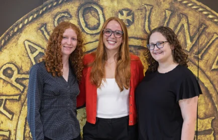 Three women, all Engineering Students from Valparaiso University and proud recipients of the Tau Beta Pi Scholarship, smiling in front of a large gold coin background, posing closely together.