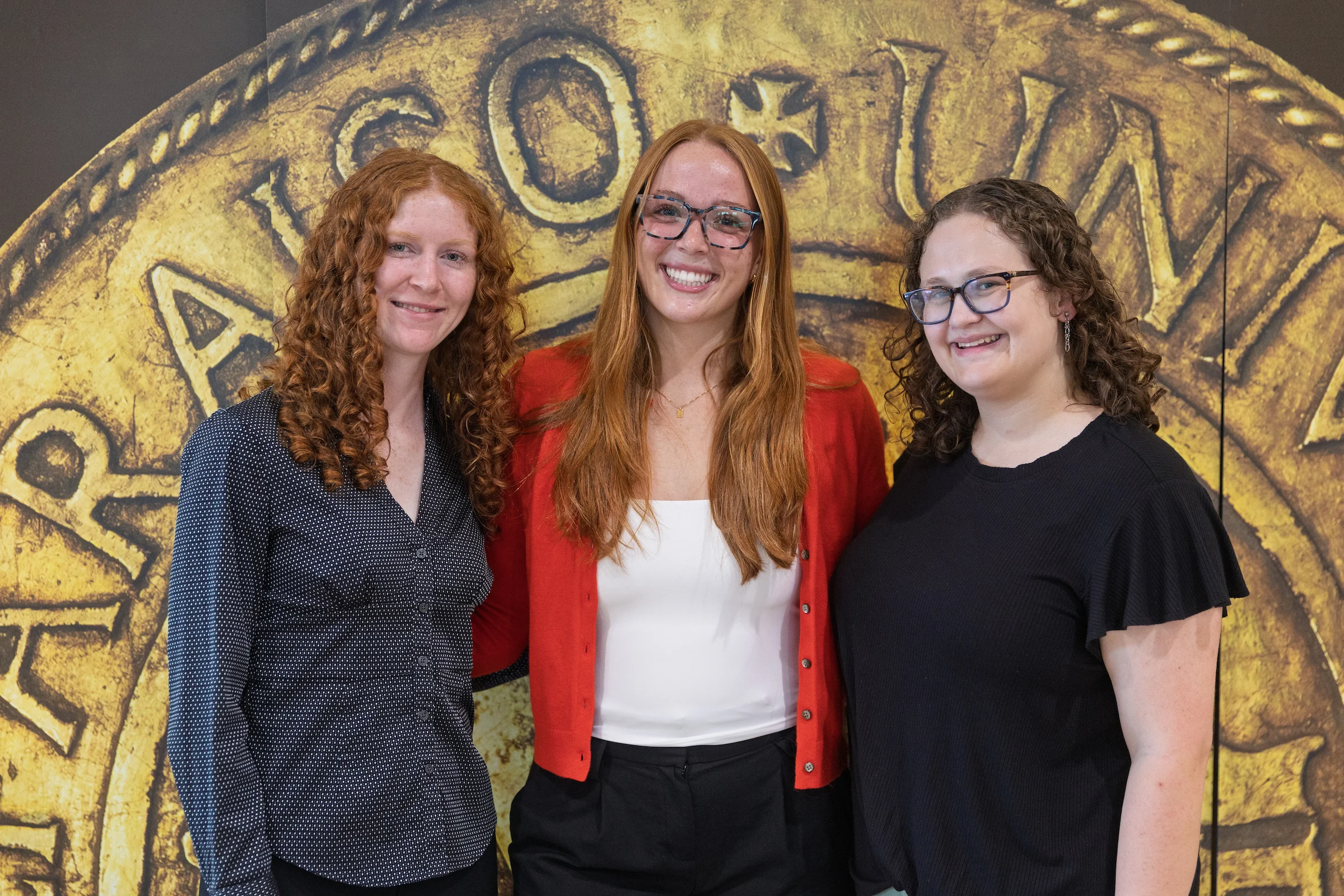 Three women, all Engineering Students from Valparaiso University and proud recipients of the Tau Beta Pi Scholarship, smiling in front of a large gold coin background, posing closely together.