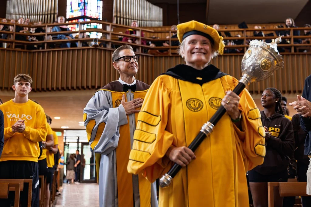 A person in a yellow academic robe holds a mace, leading the inauguration ceremony for President Rev. Brian Konkol in a church filled with people at Valparaiso University.