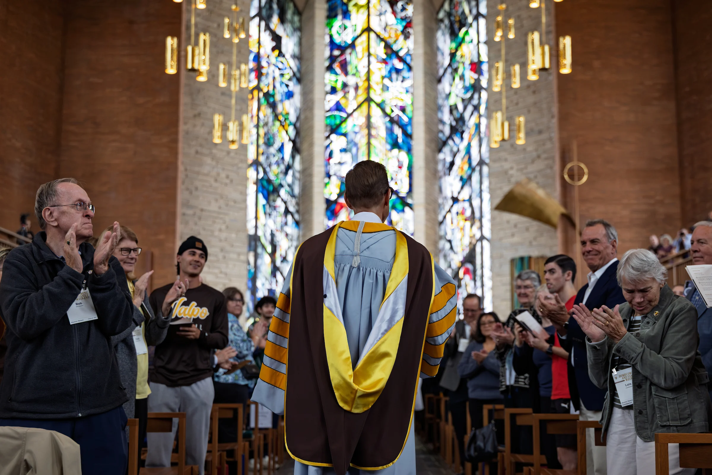 A person in academic regalia stands in a church as people around them clap and stained glass shines behind, celebrating the Inauguration of President Rev. Brian Konkol at Valparaiso University.