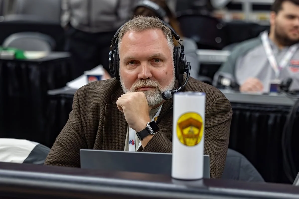 Man with headset and brown jacket sits at table with laptop, resting chin on hand, looking at camera—perhaps deep in thought as Paul Oren prepares for his next Beacon podcast episode.