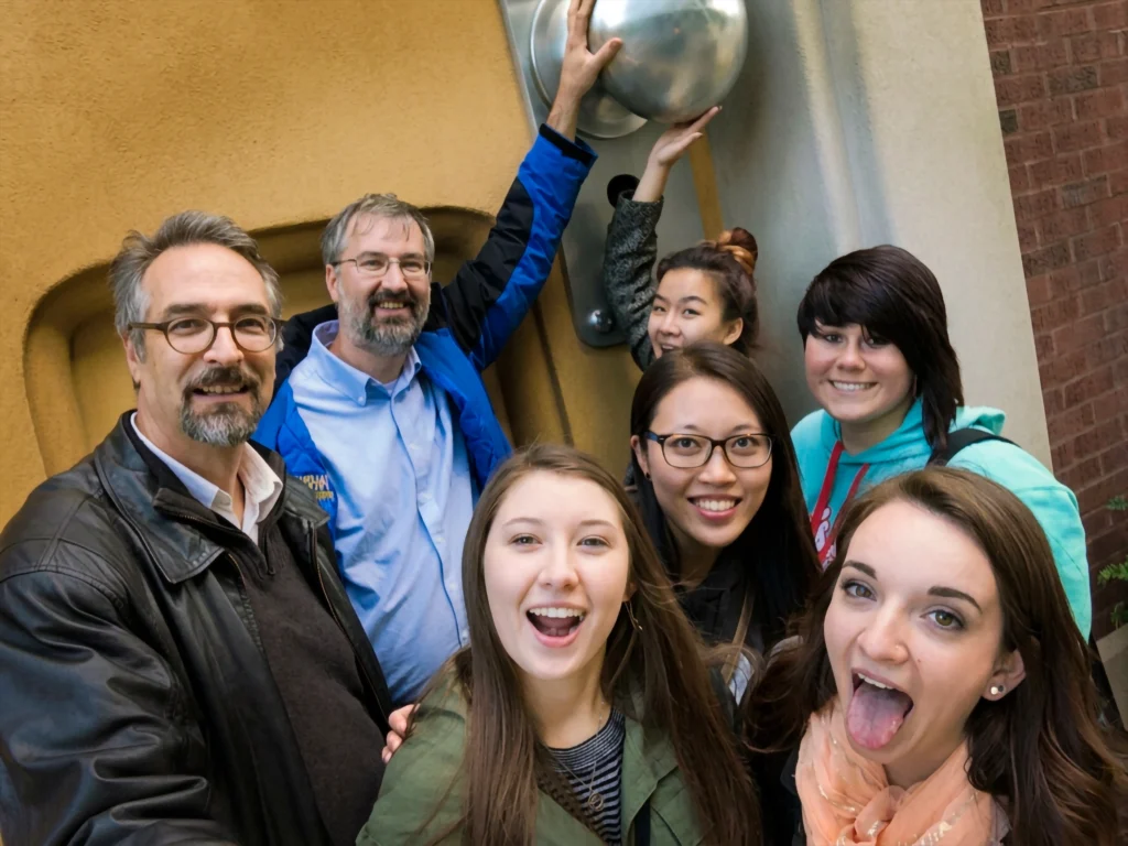 Seven people smile and pose for a selfie in front of a large yellow wall and metal sculpture.