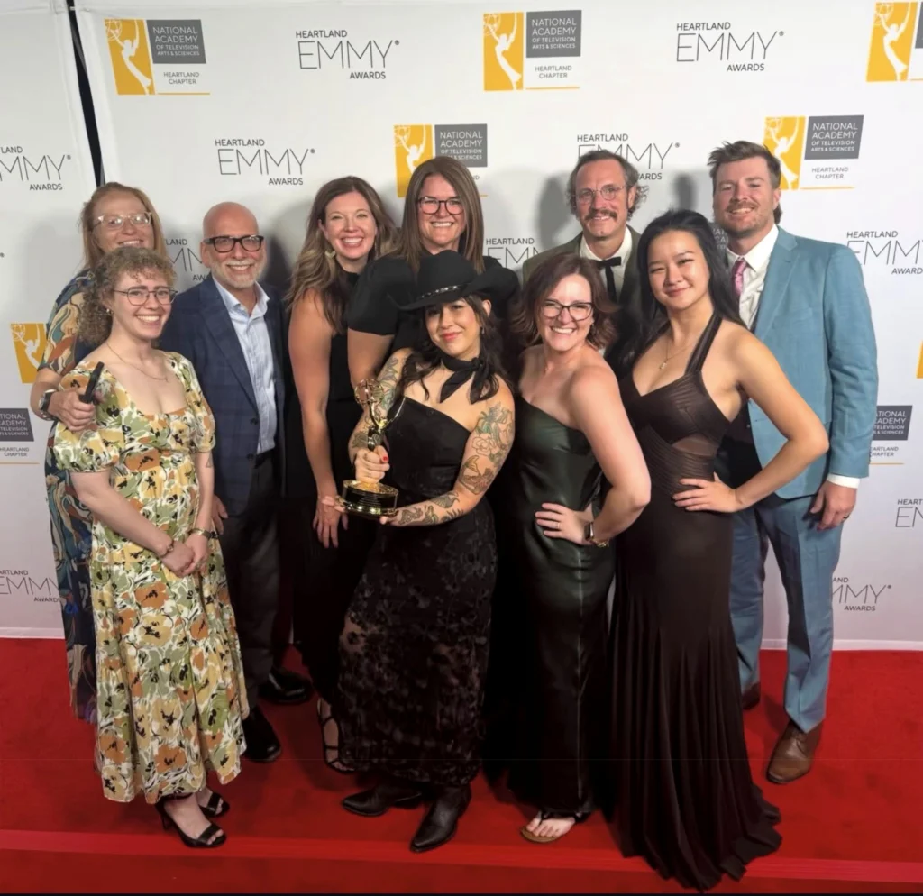 A group of people in formal wear pose on a red carpet, one holding an Emmy award.
