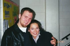 Two people smiling and posing together indoors, with a colorful poster for the Beacon, Valparaiso University’s student newspaper, in the background.