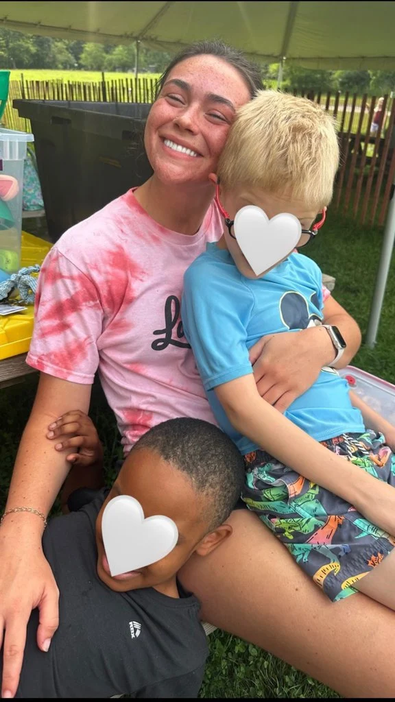 A Valparaiso University student smiles while sitting outside, holding two children with hearts covering their faces on her lap — embodying the spirit of service and purpose as a proud 2026 Valpo CAPS Fellow.