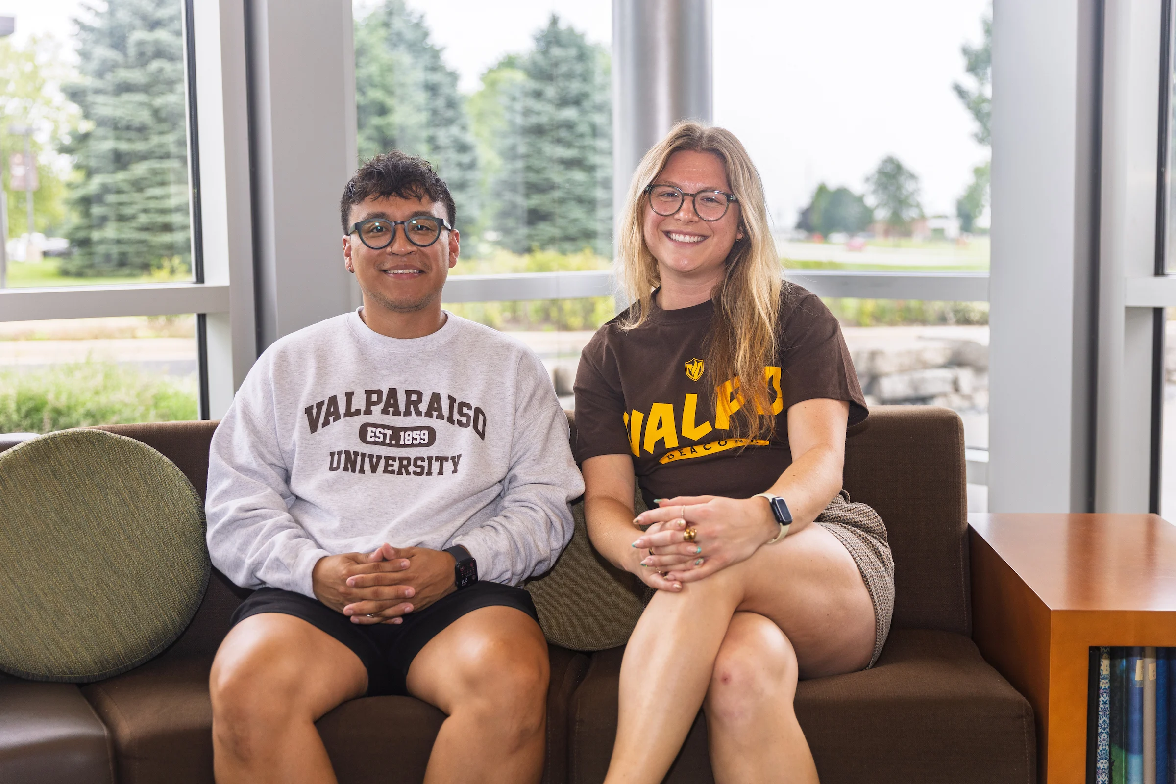 Two Valparaiso University admission counselors sit on a couch, smiling and wearing Valparaiso University shirts in a bright lounge area.