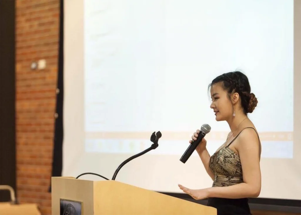 Woman holding a microphone speaks at a podium with a projector screen in the background.