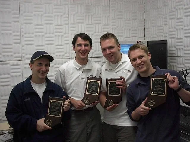 Four smiling men, including Paul Oren, stand indoors holding award plaques, posing together in a soundproofed room. Their achievements in journalism are celebrated, as highlighted by the prominent Beacon awards they proudly display.