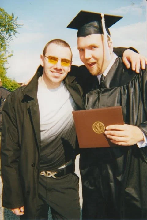 Two men smile; one is Paul Oren, wearing a graduation cap and gown holding his M.S. diploma, while the other sports sunglasses and a black coat. A sense of accomplishment shines between them, captured like a Beacon of pride.