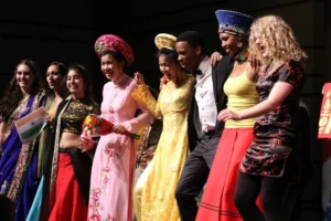 A diverse group in colorful cultural outfits stands together, smiling and linking arms on stage.