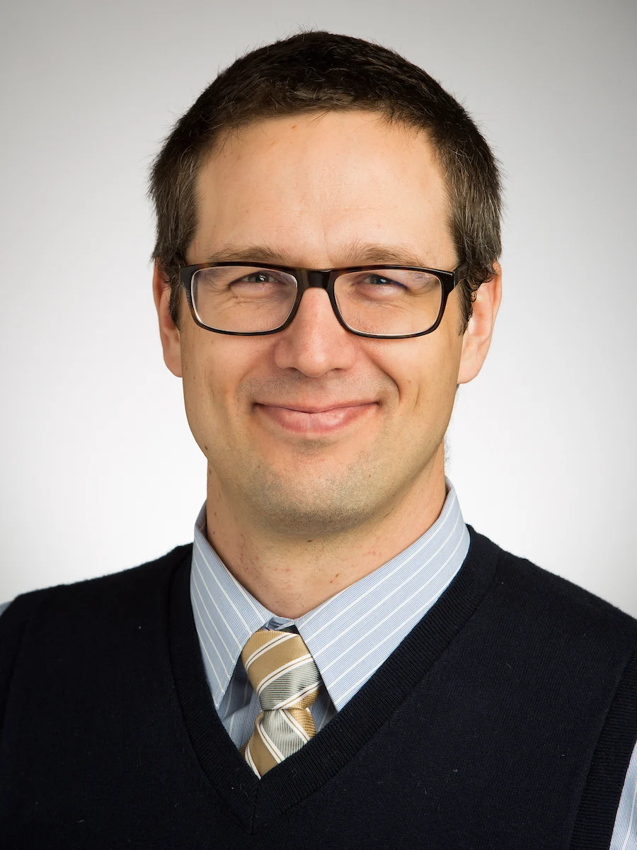 Smiling man in glasses, dress shirt, striped tie, and sweater vest for workshops.