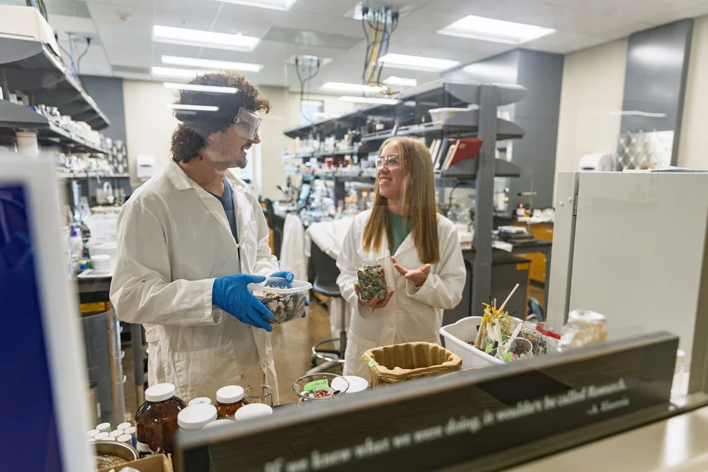 Two scientists in lab coats talk while holding containers of microplastics in a Valparaiso University laboratory.