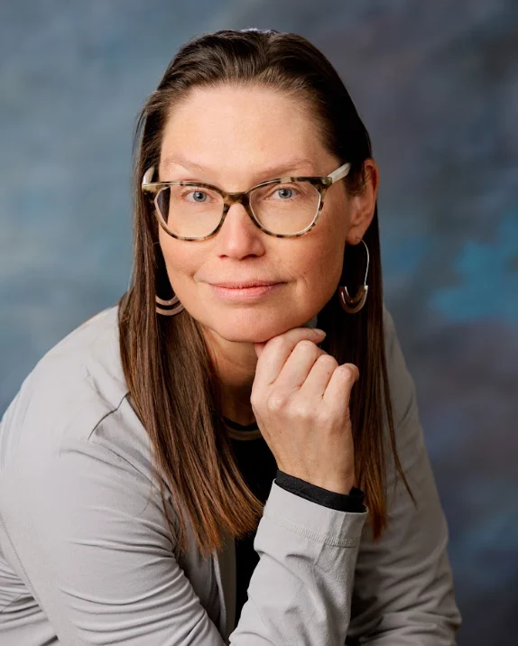 A worship leader with long brown hair, glasses, and earrings, resting her chin on her hand.