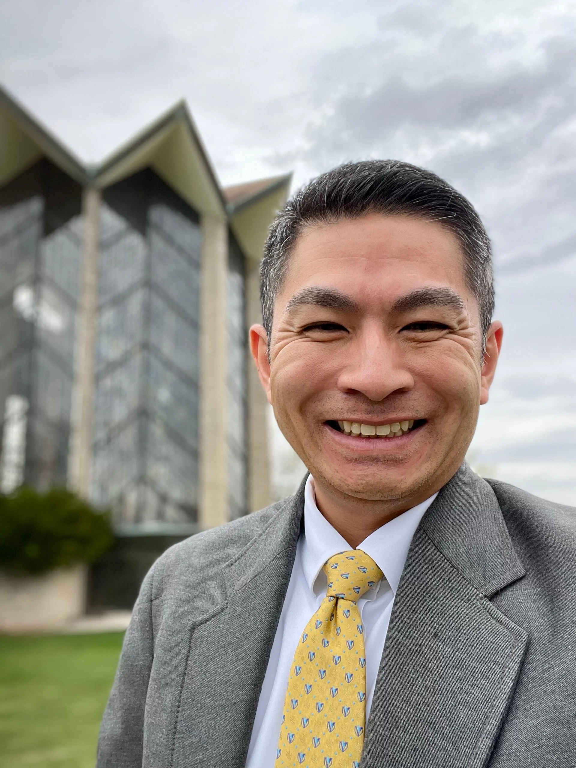 A smiling worship leader in a suit and yellow patterned tie stands in front of a modern building.