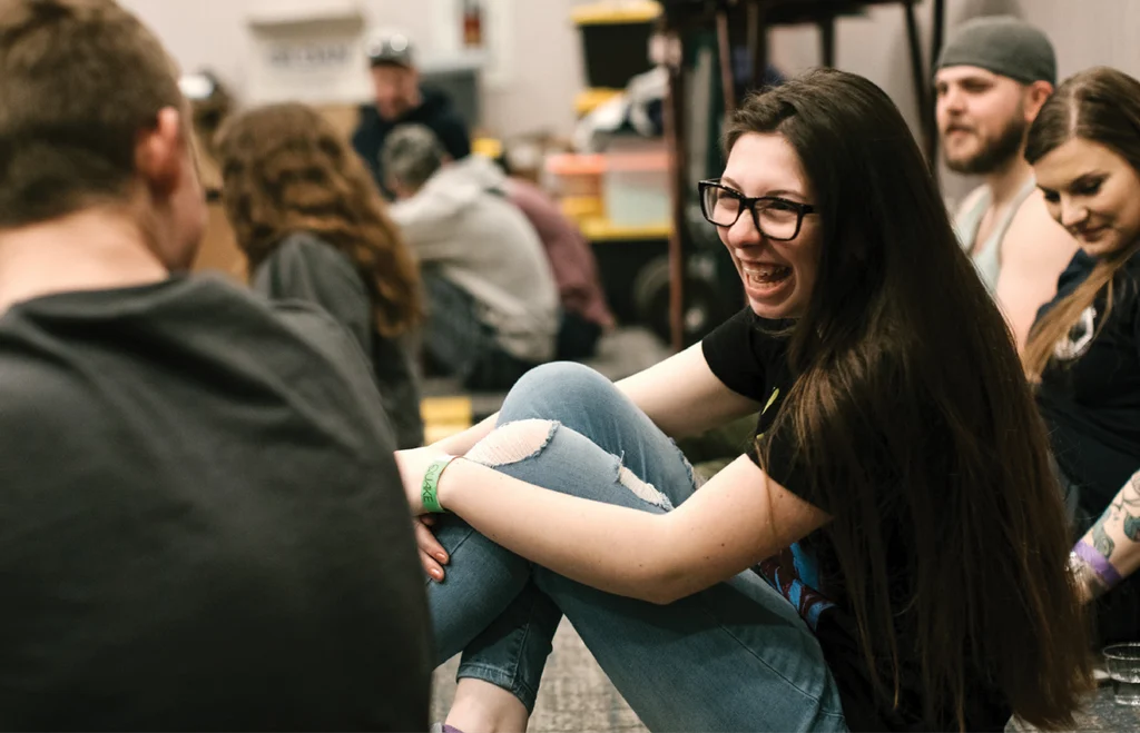 A young woman with glasses laughs while sitting in a circle with a group of people.