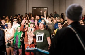 A performer on stage faces an enthusiastic crowd of young people singing along.