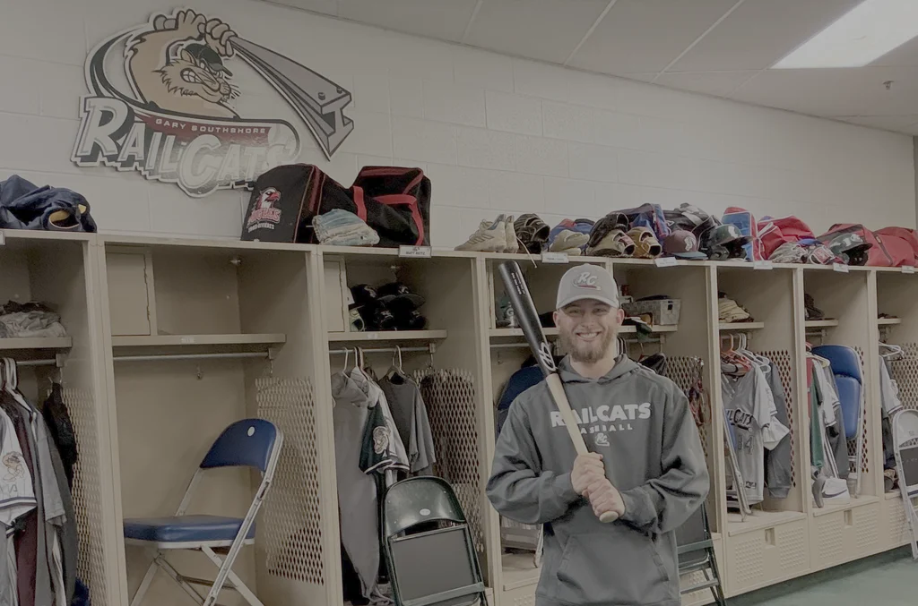 A smiling man in a Railcats hoodie holds a baseball bat in a local baseball locker room.