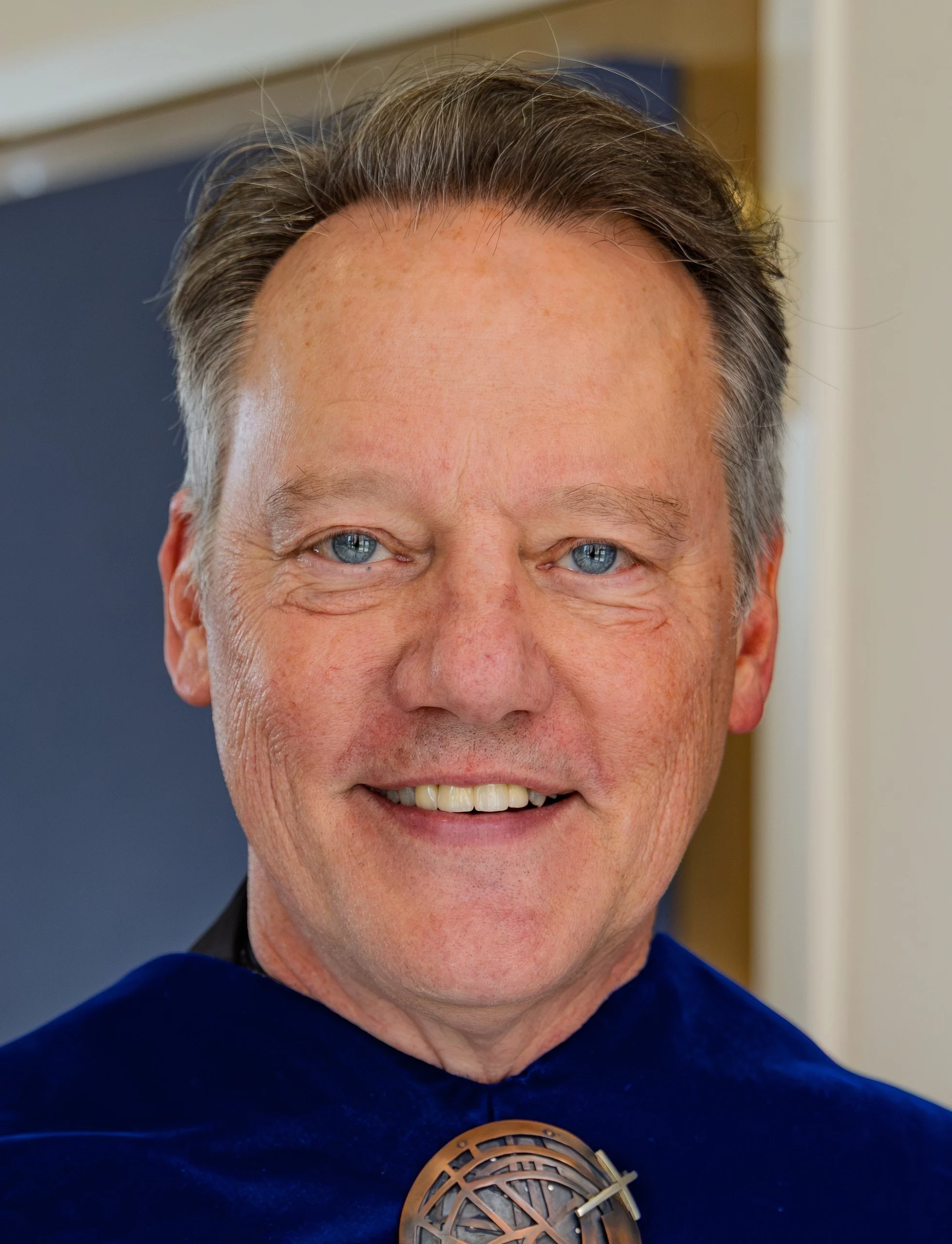 Headshot of a middle-aged man smiling in a blue academic hood with a metal emblem, captured during the plenaries.
