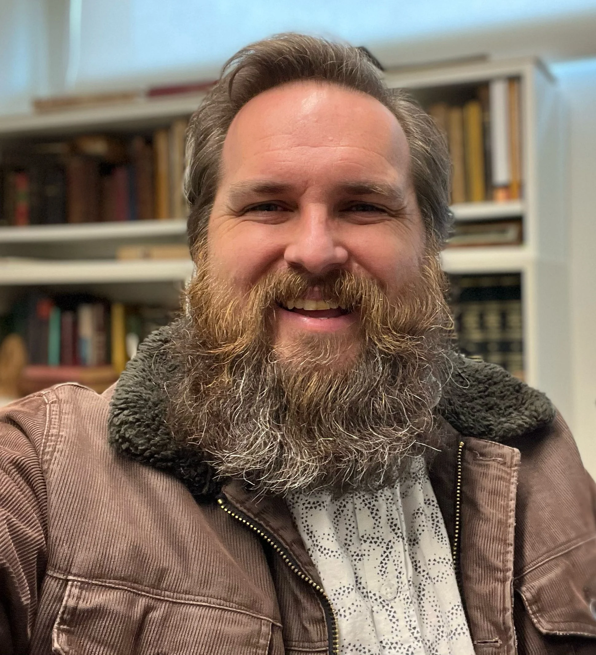 A smiling man with a full beard wearing a brown jacket with a fur-lined collar for workshops.